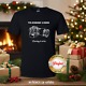 Black t-shirt with white technical drawing of camera with lens '70-200mm Zoom' above and 'Reeling it all in' below. This perfect gift is on a wood table surrounded by wrapped gifts in cozy room.