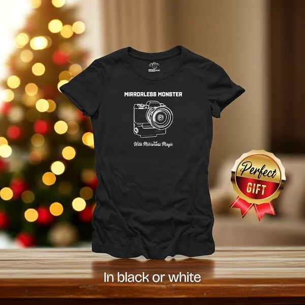 Black t-shirt with white technical line drawing of a mirrorless camera body. The text 'Mirrorless Monster' is above and 'With Mirrorless Magic' is below. The shirt is in a room with a Christmas tree, badge says 'Perfect Gift' text 'In black or white' is visible.