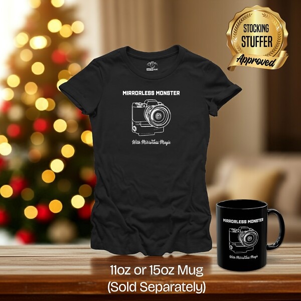 Black t-shirt and matching black mug with white technical line drawing of a mirrorless camera body. The text 'Mirrorless Monster' is above and 'With Mirrorless Magic' is below. The shirt is in a room with a Christmas tree. Stocking stuffer approved!