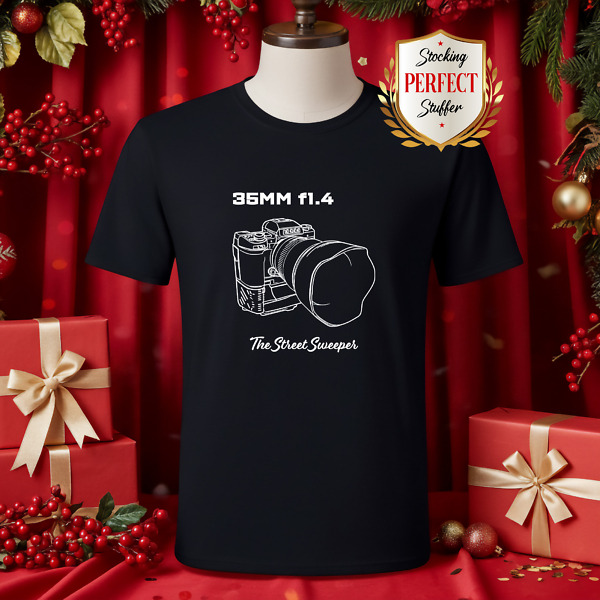 Black t-shirt with a precise line drawing of a camera with 24mm tilt shift lens, text '24MM TILT SHIFT' above and 'I'd explain, but you wouldn't understand' below. Shirt is surrounded by wrapped gifts in front of red drapery.